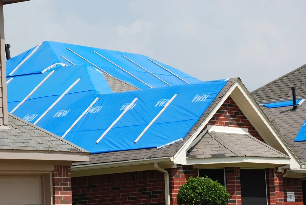 Brick home with a blue emergency tarp over storm-damaged roof shingles